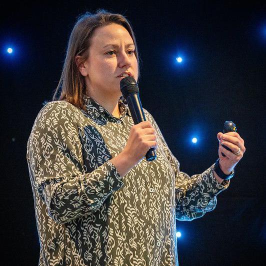 A woman holding a microphone while speaking. She is wearing a green and white shirt and has straight brown hair.