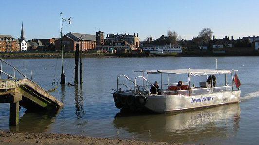 A white boat on the River Ouse. On the side of the boat it says "Lynn Ferry" in blue letters. There are two passengers and a pilot on board.