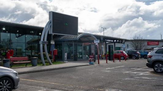 Entrance to service station, with modern single-storey building with glass frontage and curved entrance area. There are red bollards outside the entrance and cars parked in a car park.