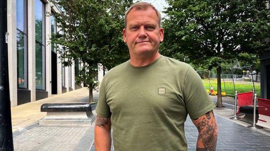 A man with tattooed arms in a green T-shirt and short grey hair faces the camera.