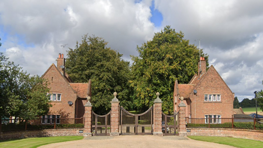 The gates at the entrance to the Chequer's estate. Three large imposing gates with red-brick houses on either side of the entrance. Before the gate is gravel, with banks of grass on either side. 