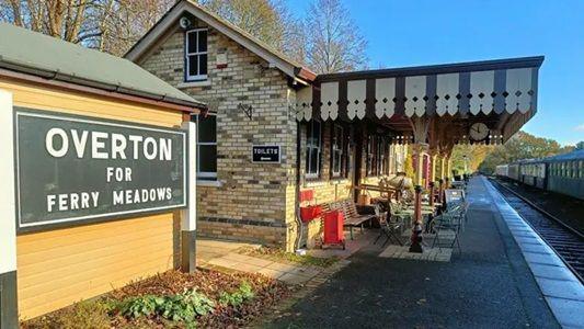 A historical railway station with a waiting room and a platform in front of it, leading to railway tracks and a black and white signboard which reads Overton for Ferry Meadows.