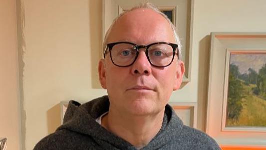 Gary Temprell is wearing a grey hooded top and dark rimmed spectacles. Behind him are a number of pictures on the wall of his home.
