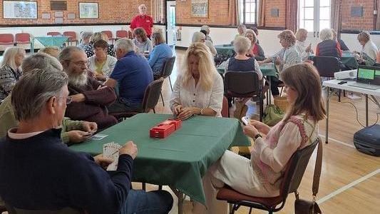 A room full of people playing bridge. There are green tablecloths on the tables and people holding cards. The room has brick walls and a wooden floor, and a man in a red top is standing behind the tables