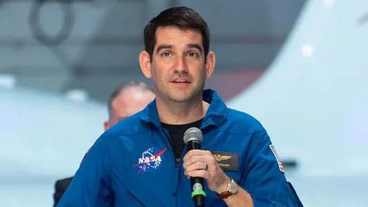 Commander Jack Hathaway giving a talk on a stage. This is a head and shoulders shot where you can see he is holding a black and silver microphone. He is looking straight ahead while wearing blue NASA overalls. There is a partial view of a man's head behind him.