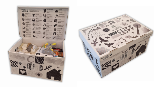 On the left is an open black-and-white patterned baby box.  Items inside include a cuddly grey rabbit with yellow ears, and white and grey socks.  Under the lid is a diagram of the box's contents. On the right is a closed box with pictures on top of nature and wildlife.