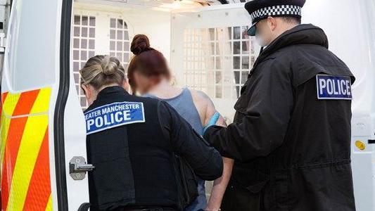 Rear view of a woman in a grey sleeveless t-shirt and brown hair tied in a bun. Under arrest, she is being led into the back of a police van by two Greater Manchester Police officers