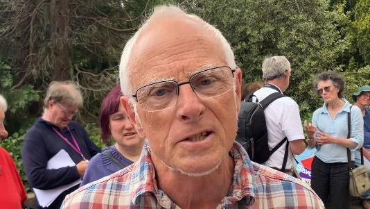 Roger Jackson, looking at the camera, at an earlier protest about cuts. He has short white hair, glasses, and is wearing a red, white and blue checked shirt. He is in the foreground of a shot with a number of other people behind him, all in front of trees.