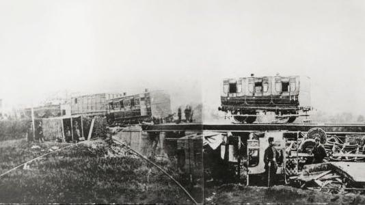 Old train carriages on a viaduct, some partially off the rails. Other carriages are destroyed underneath the viaduct in the black and white image.