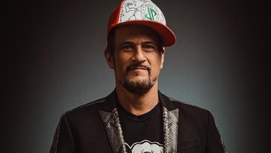 Jason Paige, wearing a dark jacket with snakeskin lapels, a dark t-shirt and a red and white baseball cap, looks into the camera.