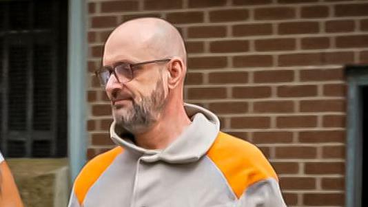 Paul Lewis outside of the court. He is wearing a grey jacket with orange on the shoulders. He is bald with a grey beard and is wearing glasses.