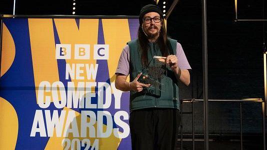 Paul Hilleard on stage with his BBC New Comedy Award trophy in 2024