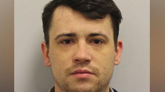 Custody image of Jordan McSweeney, a young man with dark hair wearing a dark blue sweatshirt and grey T-shirt