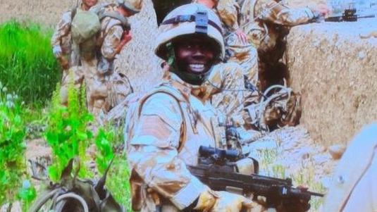 OB smiles and poses for a photo in his camouflage uniform. He holds a gun and is with a group of soldiers in the background. They are standing next to a stone wall.