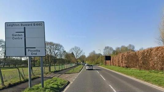 A general view of Leighton Buzzard Road in Hemel Hempstead. A white car travels in front of the camera on the left hand side of the road. There is a large white road sign on the left of the road on a grass verge. There is a pavement that runs along the left hand side, too.