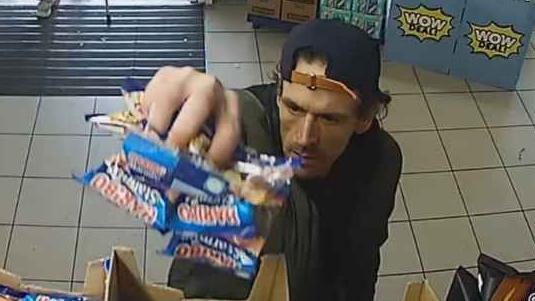A CCTV image of Aaron Kelly stealing packets of Haribos from a shop shelf.
He is a white man, wearing a cap backwards with long slightly messy hair. 