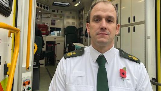 Gordon Hunt in front of an ambulance wearing a white shirt and green tie.