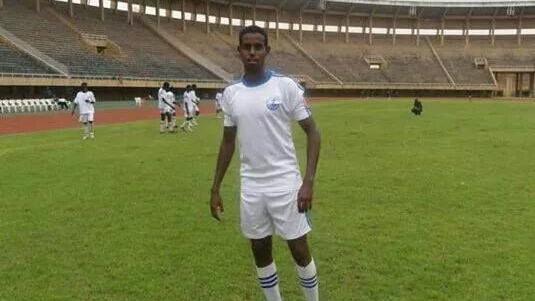 Yousuf Mohammad wearing a white football kit with blue accents in a football stadium, other players can bee seen in the background. 