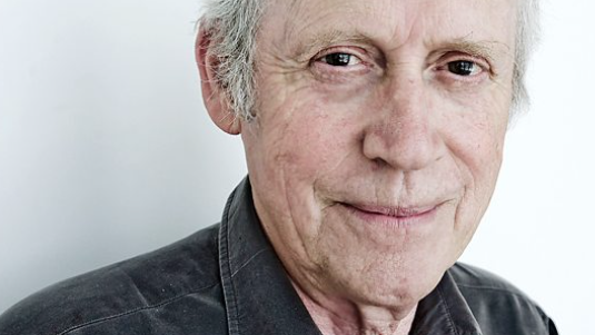 Archie Fisher is looking into the camera and half smiling. He has grey hair but the photo has been cropped at his forehead. He is wearing a grey shirt.