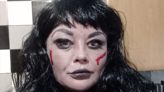 Anita has white face make-up on with red marks for blood. She is wearing a black wig.