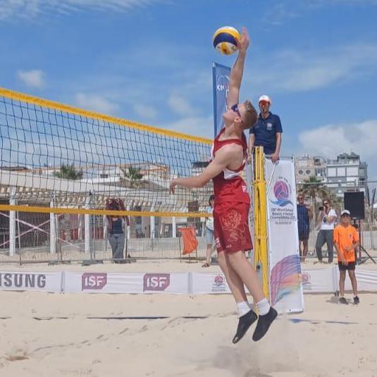 An action shot of Lucas playing beach volleyball - he is wearing a red vest and shorts and is stretching to reach the ball at the net. 