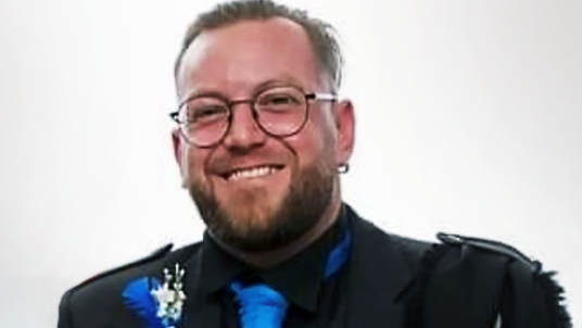 Smiling headshot of David Graham, who has a beard and is wearing glasses. He is wearing a black jacket, black shirt and a blue tie.