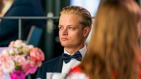 Marius Borg Høiby is wearing a black tuxedo and looking to his right while seated at a table. In the foreground, there are pink flowers on the table and a woman with her back to the camera.