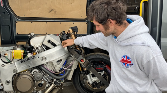 Guy Martin wearing a light grey hooded sweatshirt and leaning on his modified motorcycle.