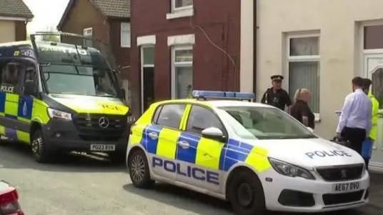 Two police cars are parked on a street outside houses. An officer in a high vis jacket speaks to a civilian woman.