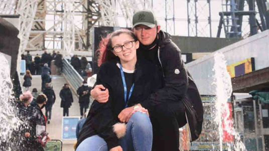 Two people are crouching together in front of a water fountain at an amusement park. They are dressed in casual clothing, including jackets and jeans, and appear to be posing for a souvenir photo. Behind them, there are visible amusement park structures.
