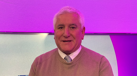 Man with a jumper looking into the camera and smiling. He has a cream jumper on with a shirt and tie. 
