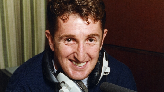 A man with curly brown haior wearing a navy fleece and smiling at the camera. He has headphones around his neck and is sat in front of a black microphone. It is a head and shoulders shot of him.
