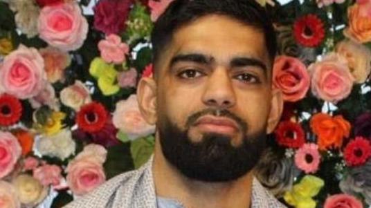 Luqman Ishfaq, a young man with dark hair and beard wearing light clothing, standing in front of a wall of flowers