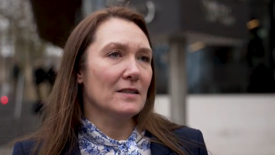 DAC Evans standing outside, with the New Scotland Yard sign blurred behind her - she is wearing a light blue and white neck scarf and a blazer and has medium length brown hair with a serious expression