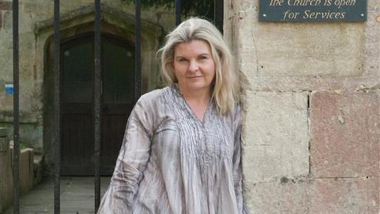 A woman smiles at the camera while resting one shoulder against a building. She has long blonde hair and is wearing a grey blouse.
