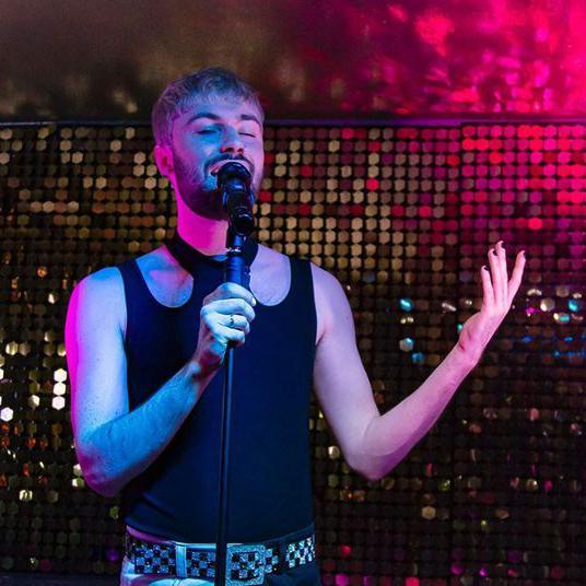 Michael T Ogilvie, wearing a tank top and singing into a micrphone, pictured against sequinned background.