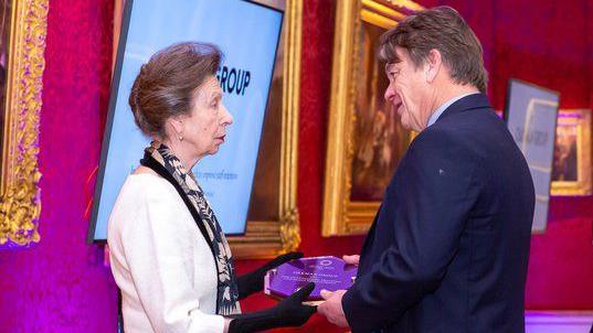Princess Anne, handing over an award to Peter Borg-Neal, at an awards event. She is wearing a white suit, with a scarf round her neck, and is wearing black gloves. Peter is wearing a dark suit and is looking at her.