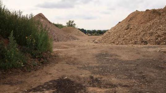 Piles of landfill on a soil-covered site. Bushes are in the foreground, and trees in the distance.