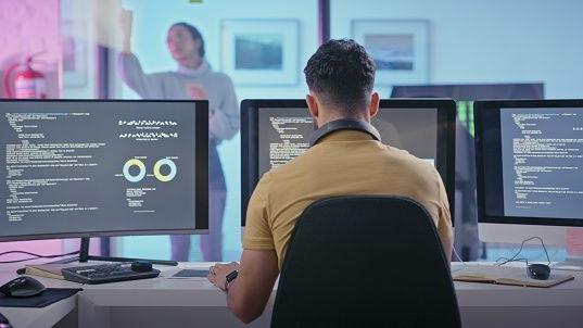 A programmer sits at their desk with multiple computer screens.