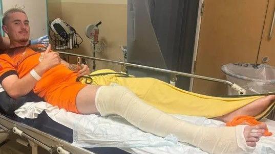 A person wearing an orange t-shirt, with his right leg in plaster, lies in a hospital bed and is giving a thumbs up gesture