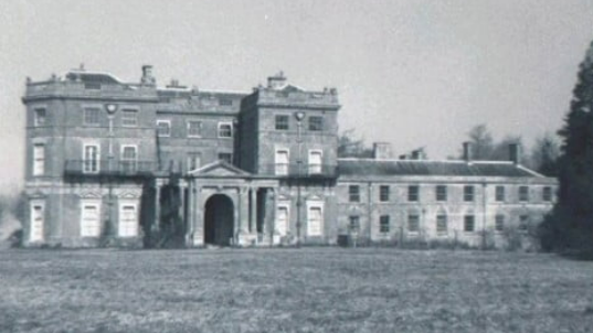 Black and white photo of a large country house, with arched entrance and considerable grounds in front of it.