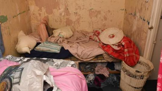 A cluttered room with a single bed. The bedding is messy and the walls are mouldy and not painted. 