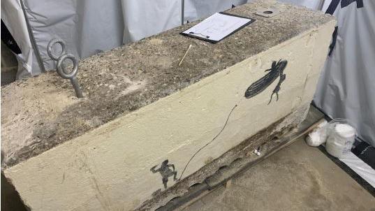 A cut out piece of concrete block with the original artwork on it. A clipboard has been placed on top of the block, which has two prongs on it.