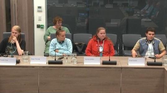 Four young people who have all spent time in care sit at a desk with microphones in front of them. They are dressed casually and all have headphones around their necks. 