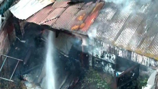 A large outhouse building in Workington which has corrugated roofing and looks decrepit has smoke coming out of the top. There is a jet of water from a fire service hose being directed into the shed.