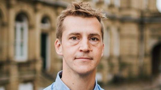 Calderdale's Labour MP Josh Fenton-Glynn - a white man with blonde hair and a blue shirt standing in front of sandstone buildings