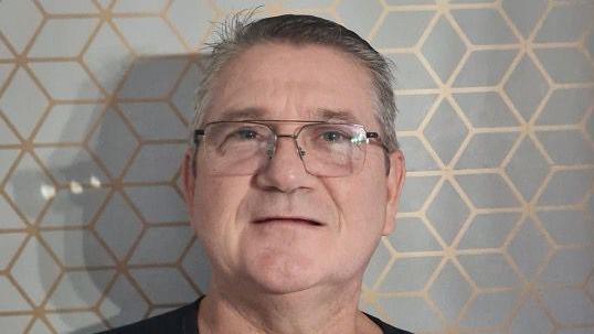 A man with short grey hair stands in front of a grey and gold patterned wallpaper. He has large glasses and is clean shaven. 