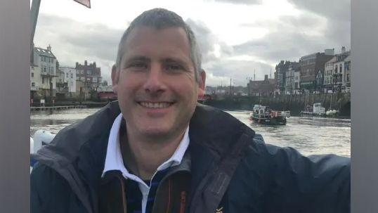 A man with short grey hair, wearing a blue shirt, brown fleece and navy jacket, sits on a boat. A canal can be seen behind him with tall buildings.