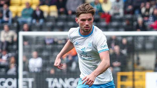 Joel Tabiner, in a white Crewe away top, looks down at the ball during a League Two game before his injury