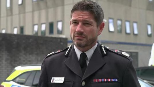 A man with brown hair and a short beard in a police uniform. Part of a police car and a concrete building is visible in the background.